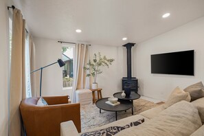 A living room with a sofa, a coffee table, a TV, a fireplace, and a window with curtains. - West House - Minutes To Autzen 2 Bedroom Home (Springfield)
