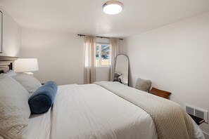 A hotel room with a large bed, a bedside table with a lamp, a mirror, and a window with curtains. - West House - Minutes To Autzen 2 Bedroom Home (Springfield)