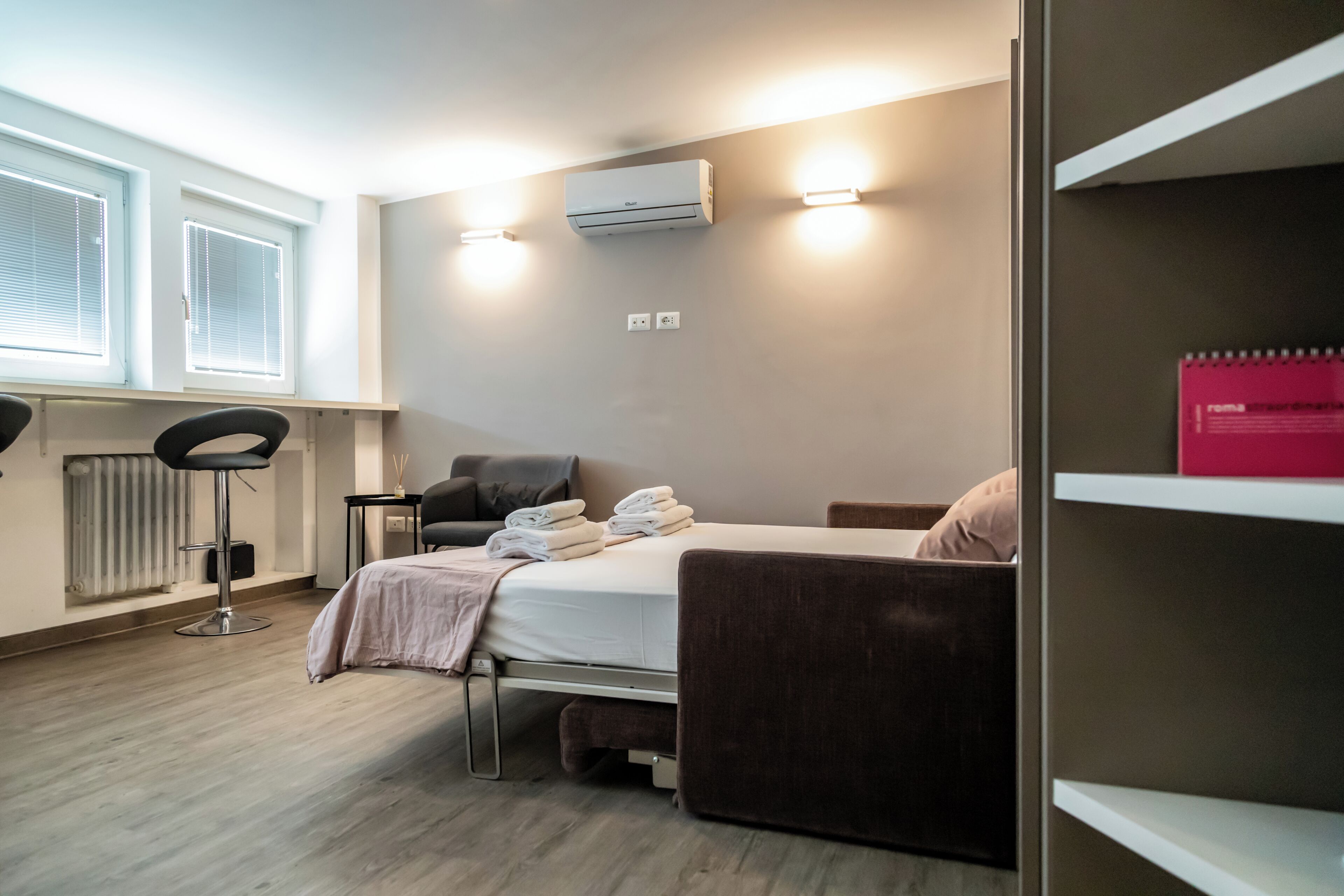 A hotel room with a bed, a chair, a desk, a radiator, and a window with blinds.