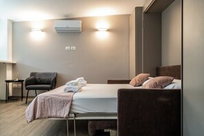 A modern hotel room with a bed, bedside tables, a chair, an air conditioning unit, and a wall-mounted light fixture. - Circo Massimo Room  (Roma)