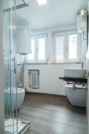 Un bany modern amb una cabina de dutxa de vidre, un lavabo blanc i un vàter. Hi ha finestres amb persianes i un radiador a la paret.