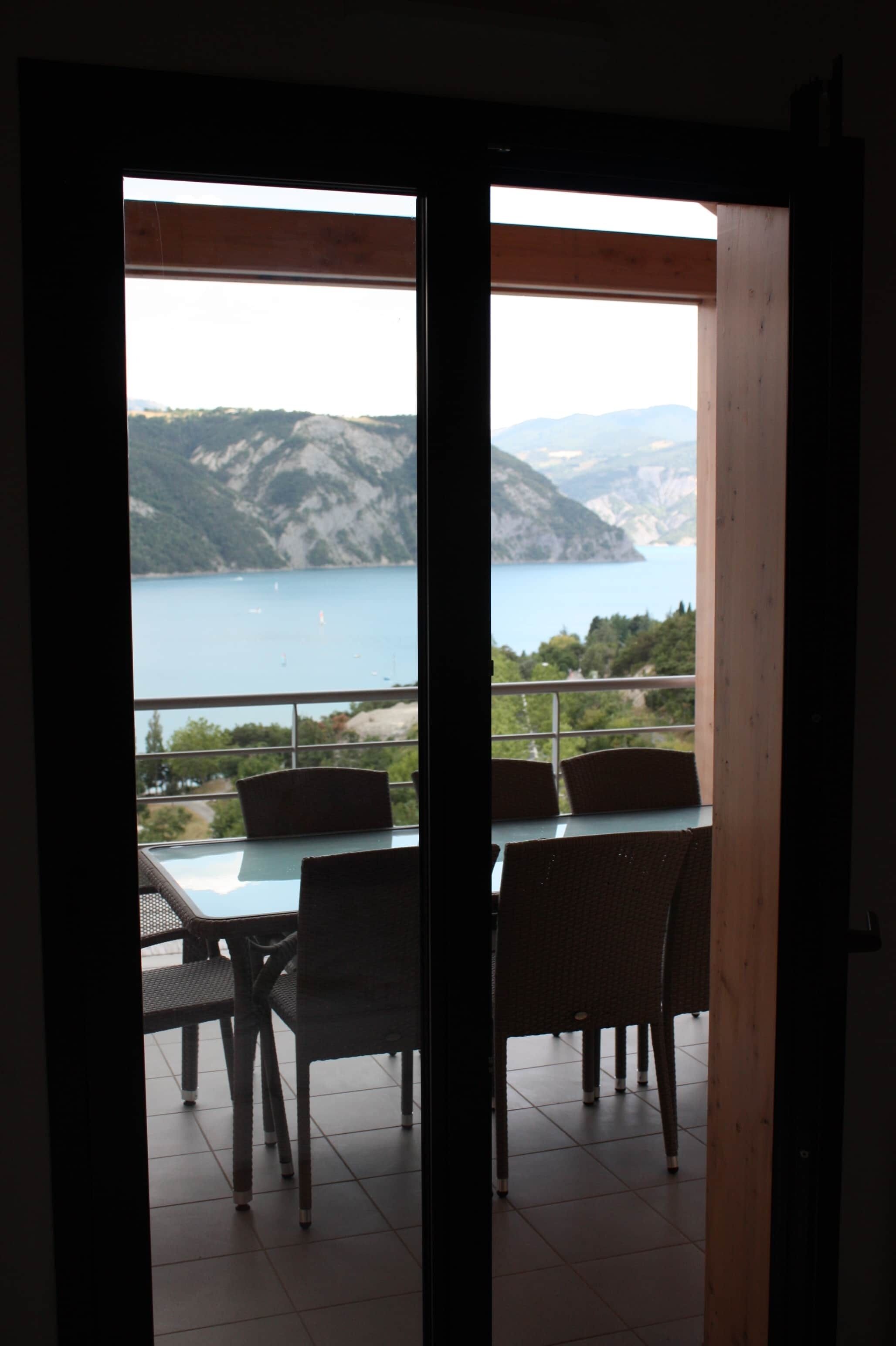 An open door leading to a balcony with a table and chairs, overlooking a body of water and hills.