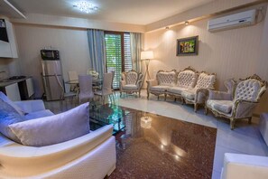 Ein Wohnzimmer mit Glastisch, weißen Sofas, einem Essbereich mit Glastisch sowie einer Küche mit Mikrowelle und Kühlschrank.