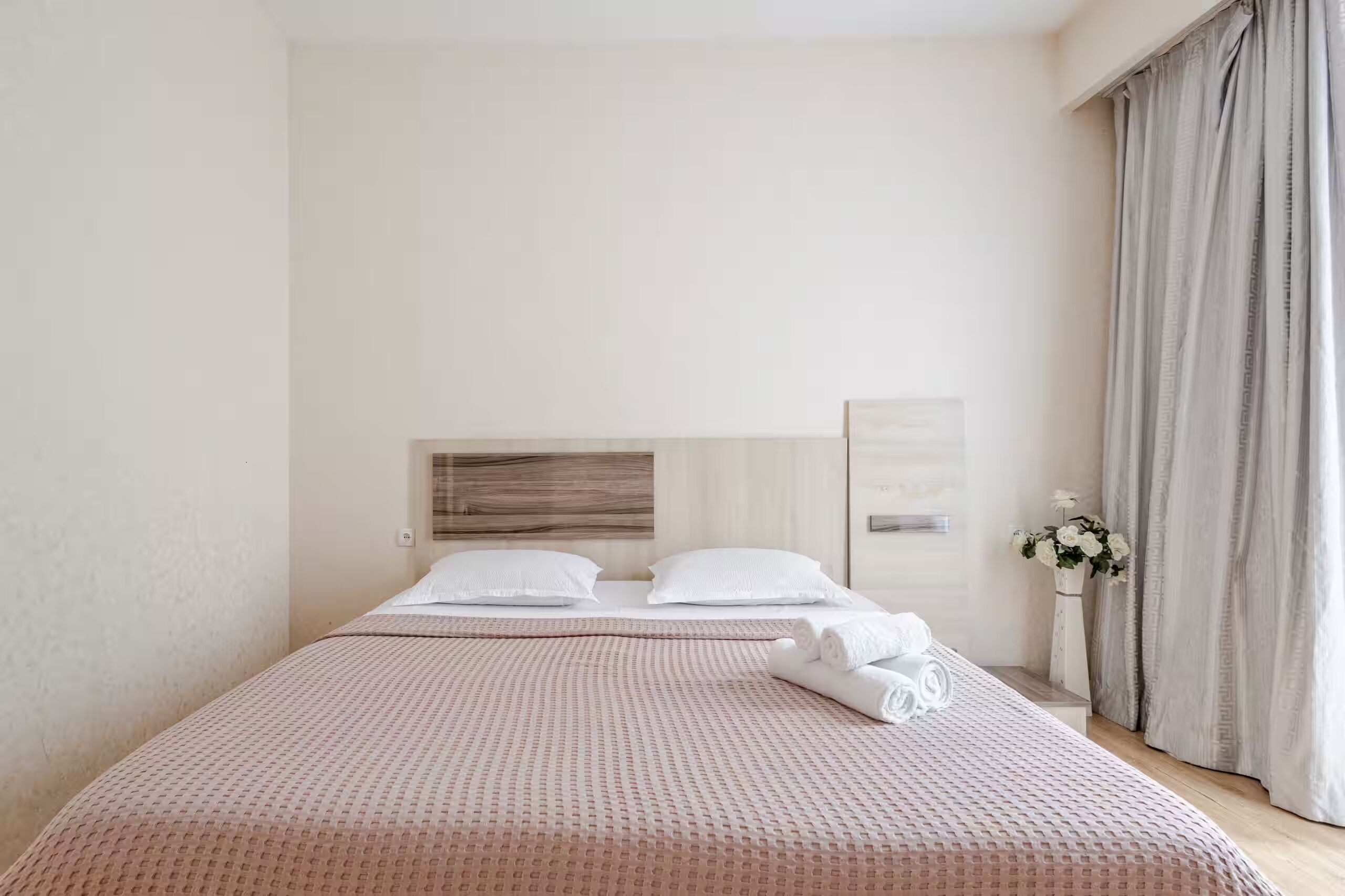 A hotel room with a bed, a headboard, a vase with flowers, and a window with curtains.