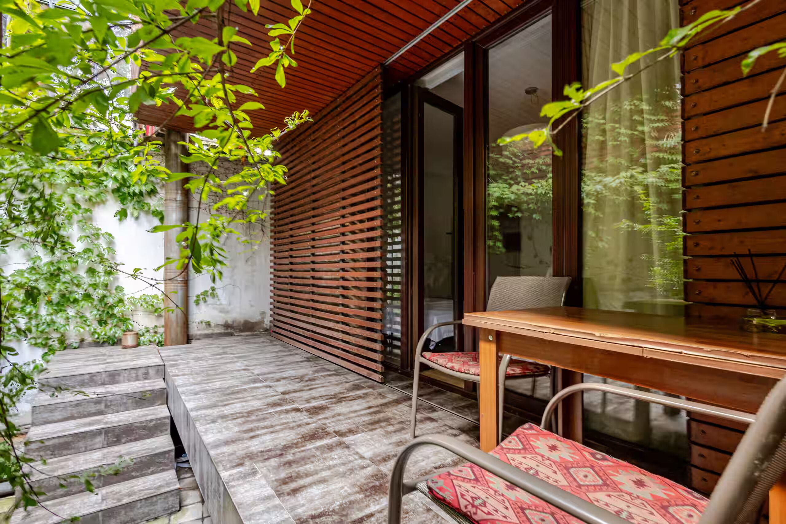 Um deck de madeira com mesa e cadeiras, uma porta de vidro e uma escadaria que leva a um andar superior.