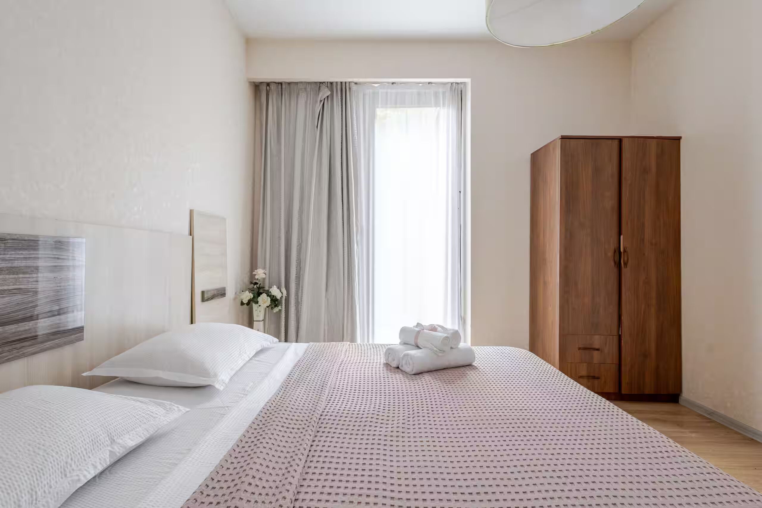 Hotellihuone, jossa on sänky, vaatekaappi, ikkuna verhoilla ja seinälle ripustettu taideteos.