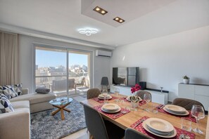 A modern living room with a dining area, large windows, and a city view. - Xag ra Apartment With Sea Views Balcony Gozo (Xaghra)