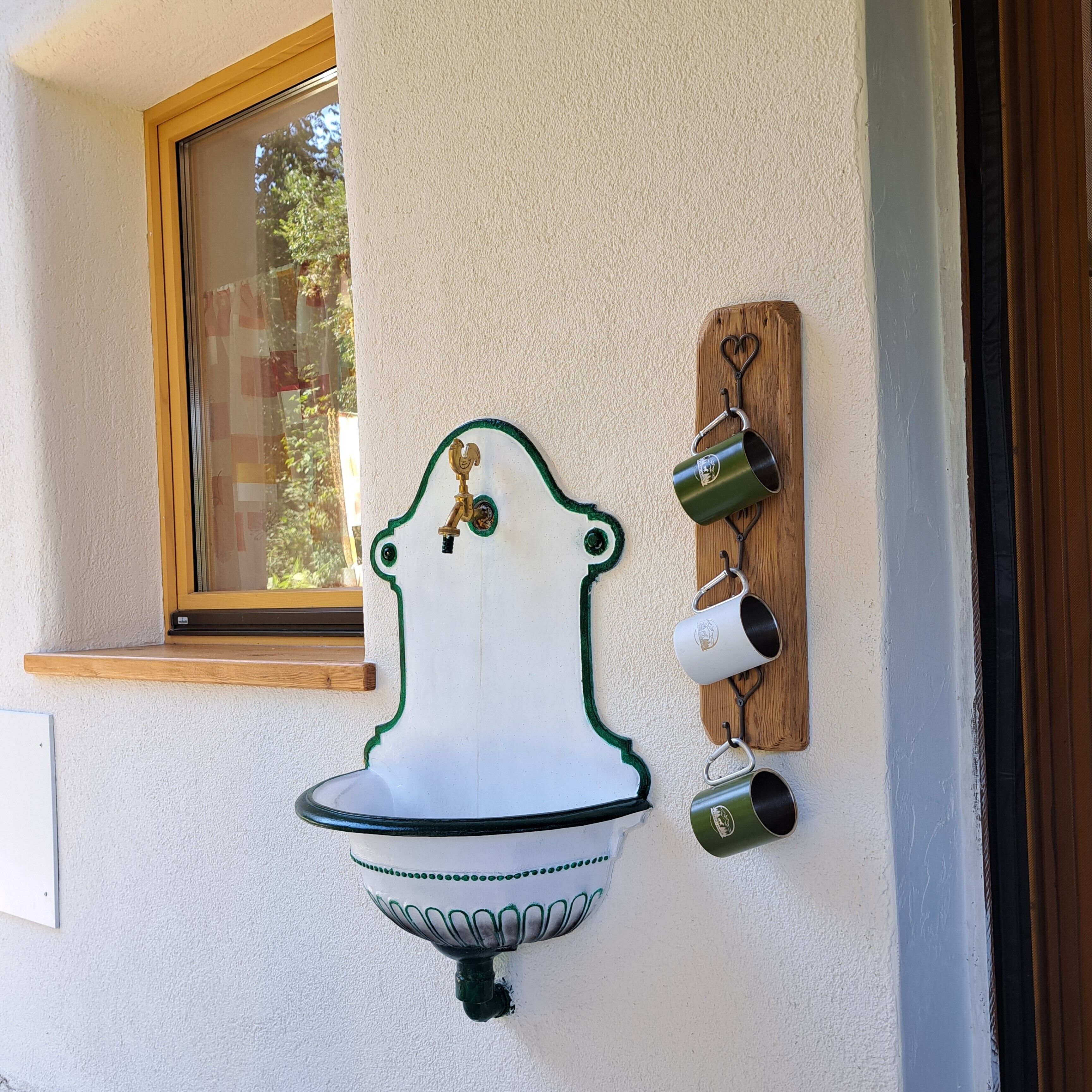 A wall-mounted outdoor sink with a decorative design, a window above it, and a wooden board with hanging mugs.