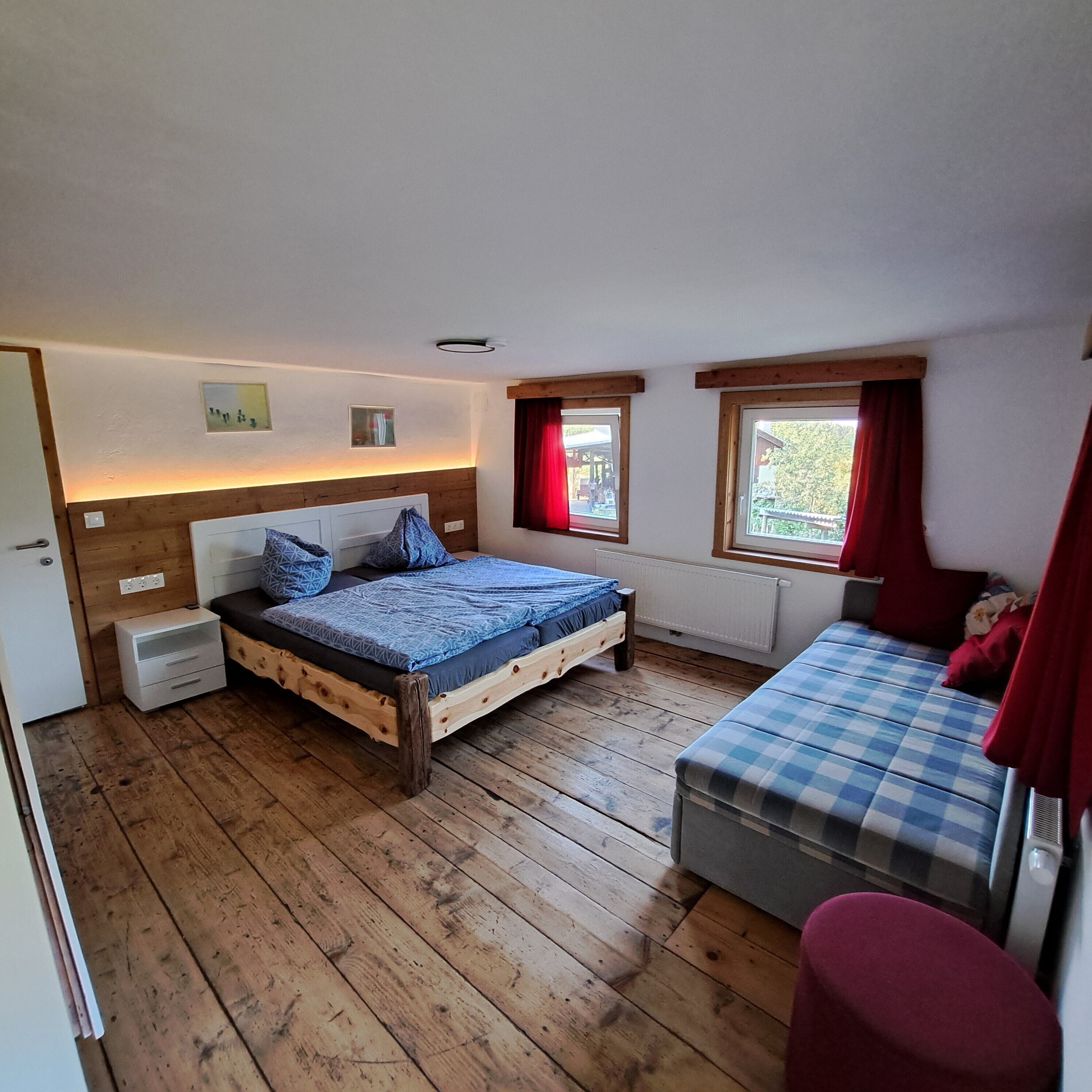 Una camera da letto con pavimento in legno, un letto con biancheria blu, un comodino, una panca con rivestimento a scacchi e due finestre con tende rosse.