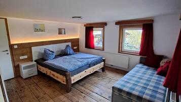 Una camera da letto con pavimento in legno, un letto con biancheria blu, un comodino, una panca con rivestimento a scacchi e due finestre con tende rosse.