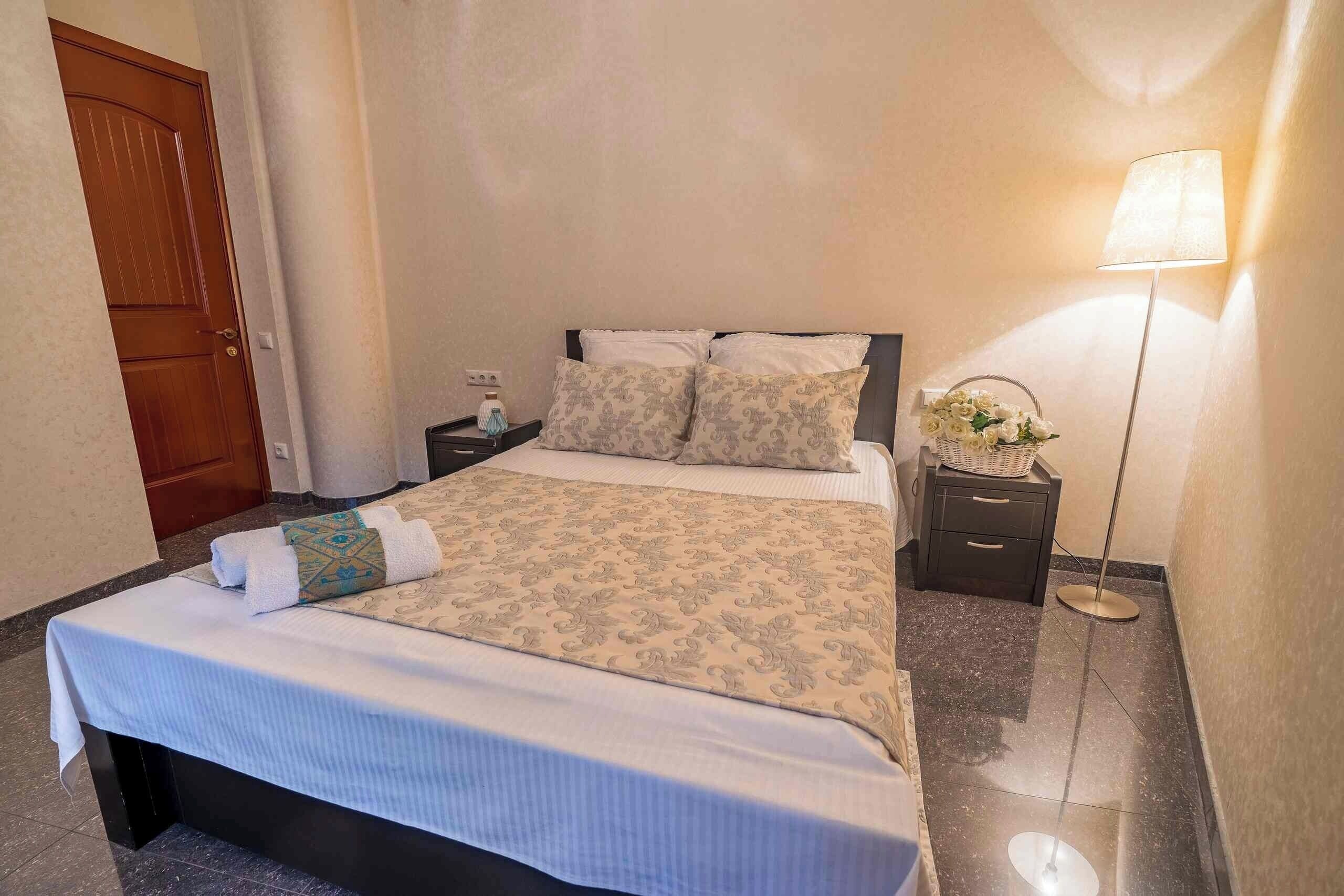 Una cama bien hecha con una colcha estampada, una mesita de noche con una lámpara y una puerta al fondo.