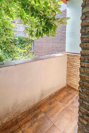 Un balcón con piso de baldosas, barandilla de madera y vista a un muro de ladrillo y follaje.