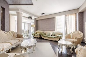 Amplia sala de estar con muebles ornamentados, una mesa de centro de mármol y ventanas grandes con cortinas.