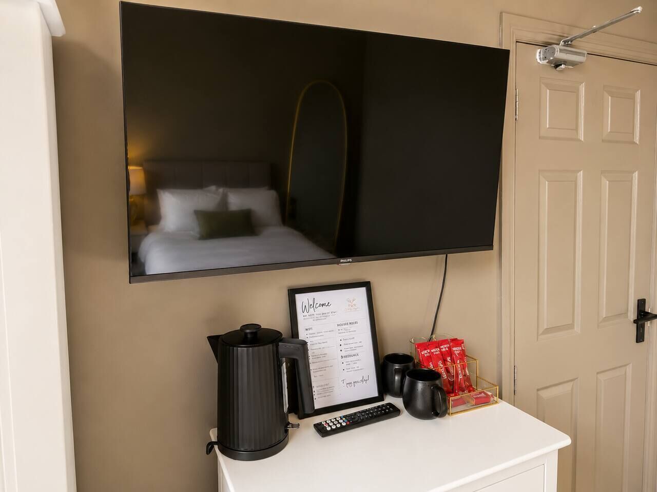 A hotel room with a bed, a television mounted on the wall, a coffee maker, a framed welcome sign, and a door.