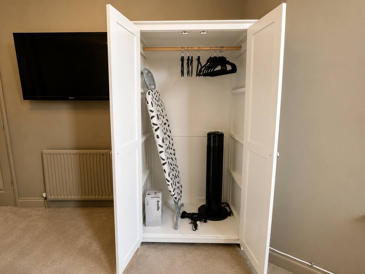 A walk-in closet with a hanging rack, ironing board, and a black cylindrical object.