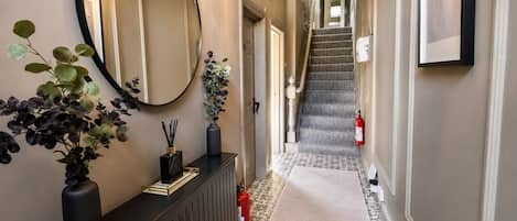 A hallway with a radiator, a round mirror, a potted plant, a fire extinguisher, and a staircase leading to another floor.