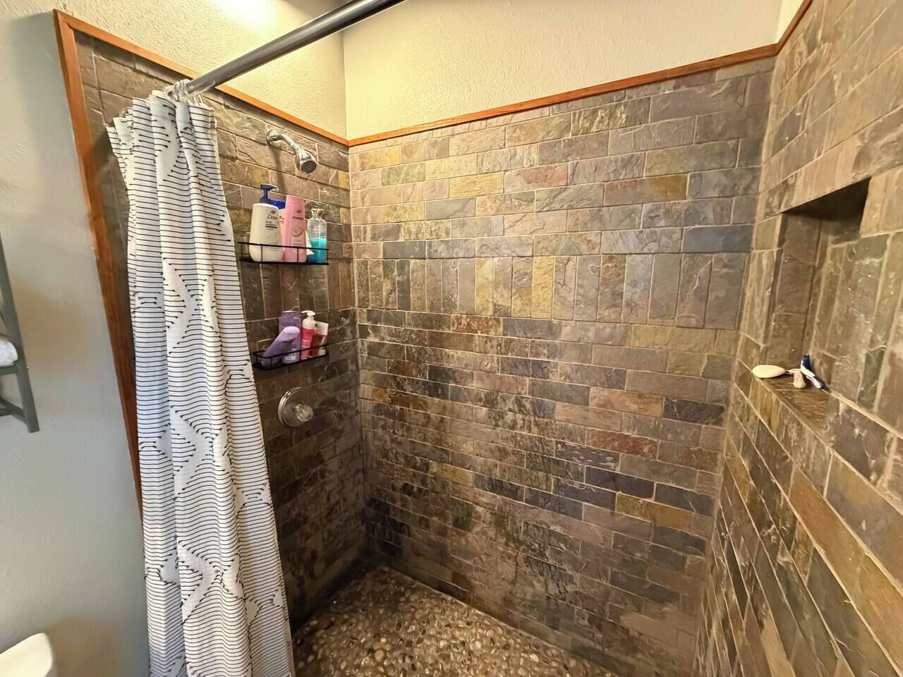 A shower area with a stone wall, a shower curtain, and toiletries on a shelf.