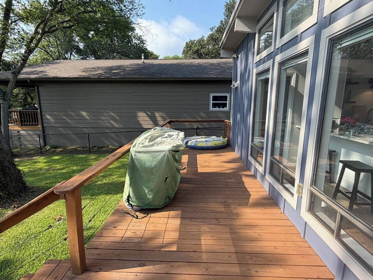 Une terrasse en bois dotée d’un espace salon couvert et d’un barbecue abrité, située à côté d’un bâtiment avec de grandes fenêtres.