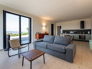 Ein modernes Wohnzimmer mit Sofa, Couchtisch und Stuhl. Es gibt eine Küchenzeile mit Esstisch und Blick ins Freie.