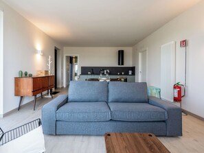 Ein modernes Wohnzimmer mit einem grauen Sofa, einem Holztisch und einer Küche im Hintergrund.