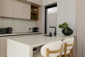 A modern kitchen with a central island, two bar stools, a coffee machine, and a large green plant. - Botanica Pru Jampa New 3BR Pool Villa (Thep Krasatti)