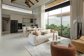 A modern living room with a sofa, coffee table, and chairs, a dining area with a table and chairs, and a kitchen in the background.