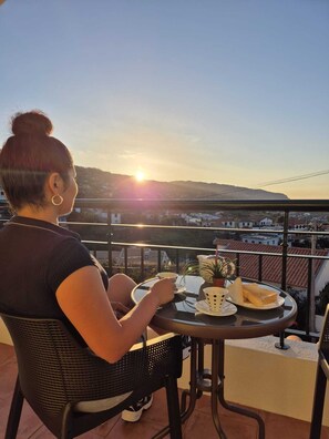A person seated at a table on a balcony overlooking a cityscape during sunset. - Cozy Two-Bedroom Attic Retreat: Rooftop, Ocean, and Sunrise Views (Ponta do sol)
