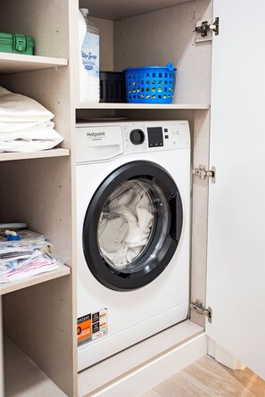 A compact washing machine in a small laundry room with a shelf above containing folded clothes and a blue laundry basket. - Cozy Two-Bedroom Attic Retreat: Rooftop, Ocean, and Sunrise Views (Ponta do sol)