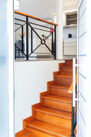 A wooden staircase with a metal railing leading to a room with a kitchen area. - Cozy Two-Bedroom Attic Retreat: Rooftop, Ocean, and Sunrise Views (Ponta do sol)