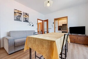 Ein Wohnzimmer mit einer Couch, einem Esstisch mit Tischdecke, einem Fernseher und einem durchgehenden Türblick in den Küchenbereich.