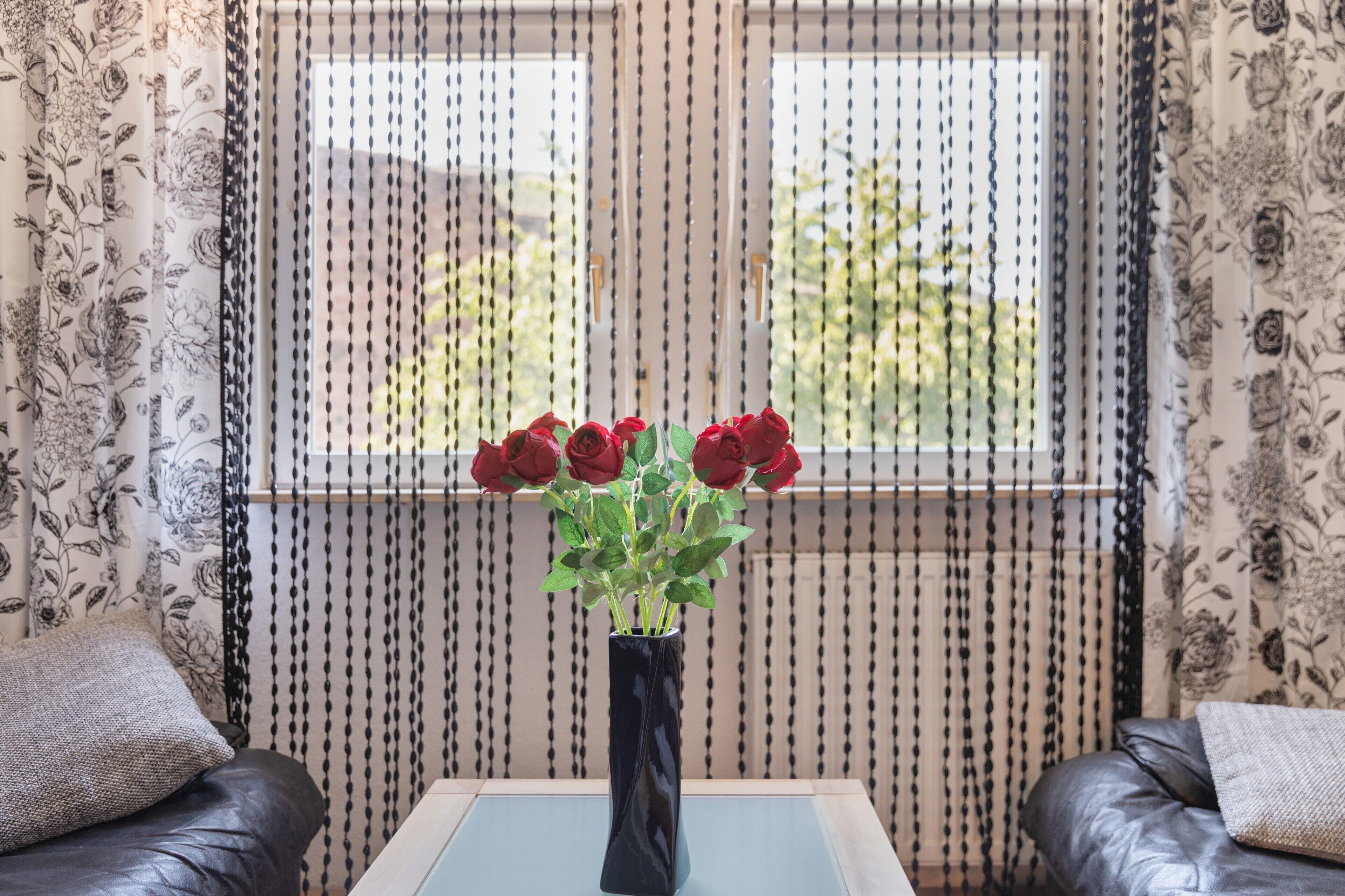A room with a floral-patterned curtain, a vase with red roses, a glass coffee table, and two leather sofas.