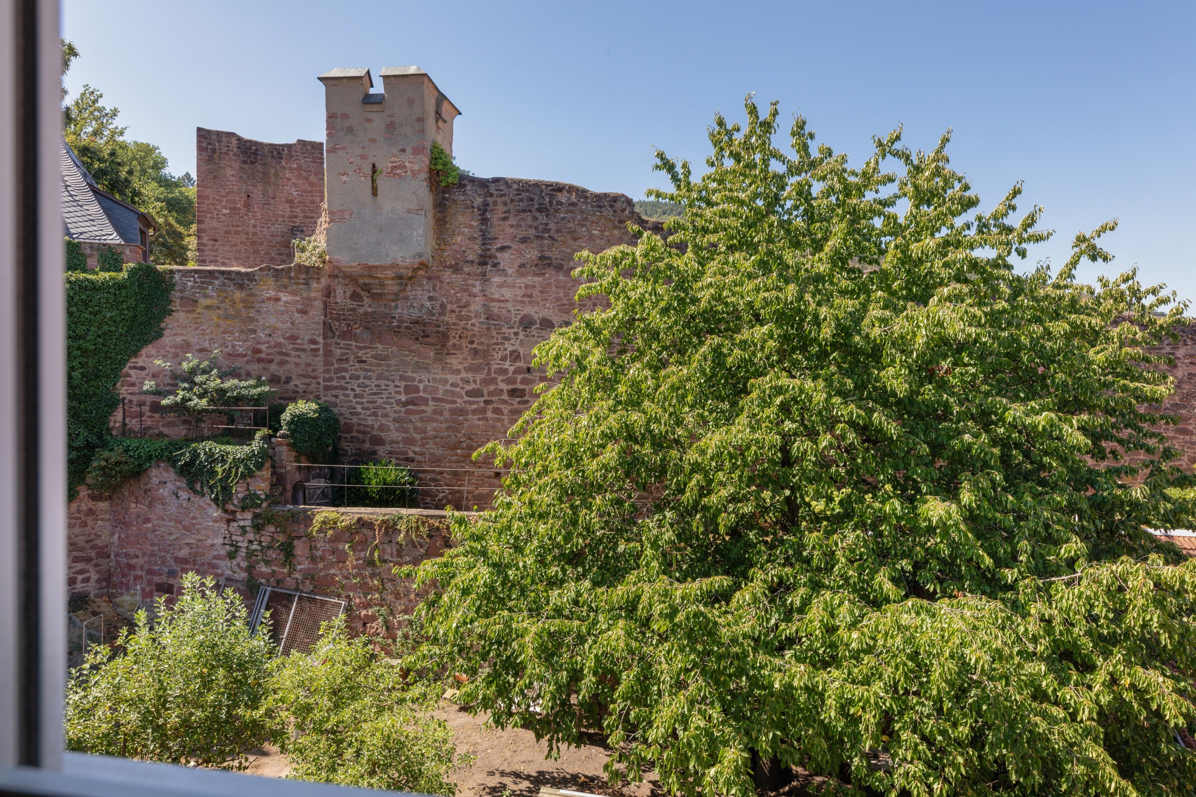 Un mur de pierre ancien, surmonté d’une petite tour, entouré de verdure et délimité par une clôture en métal.