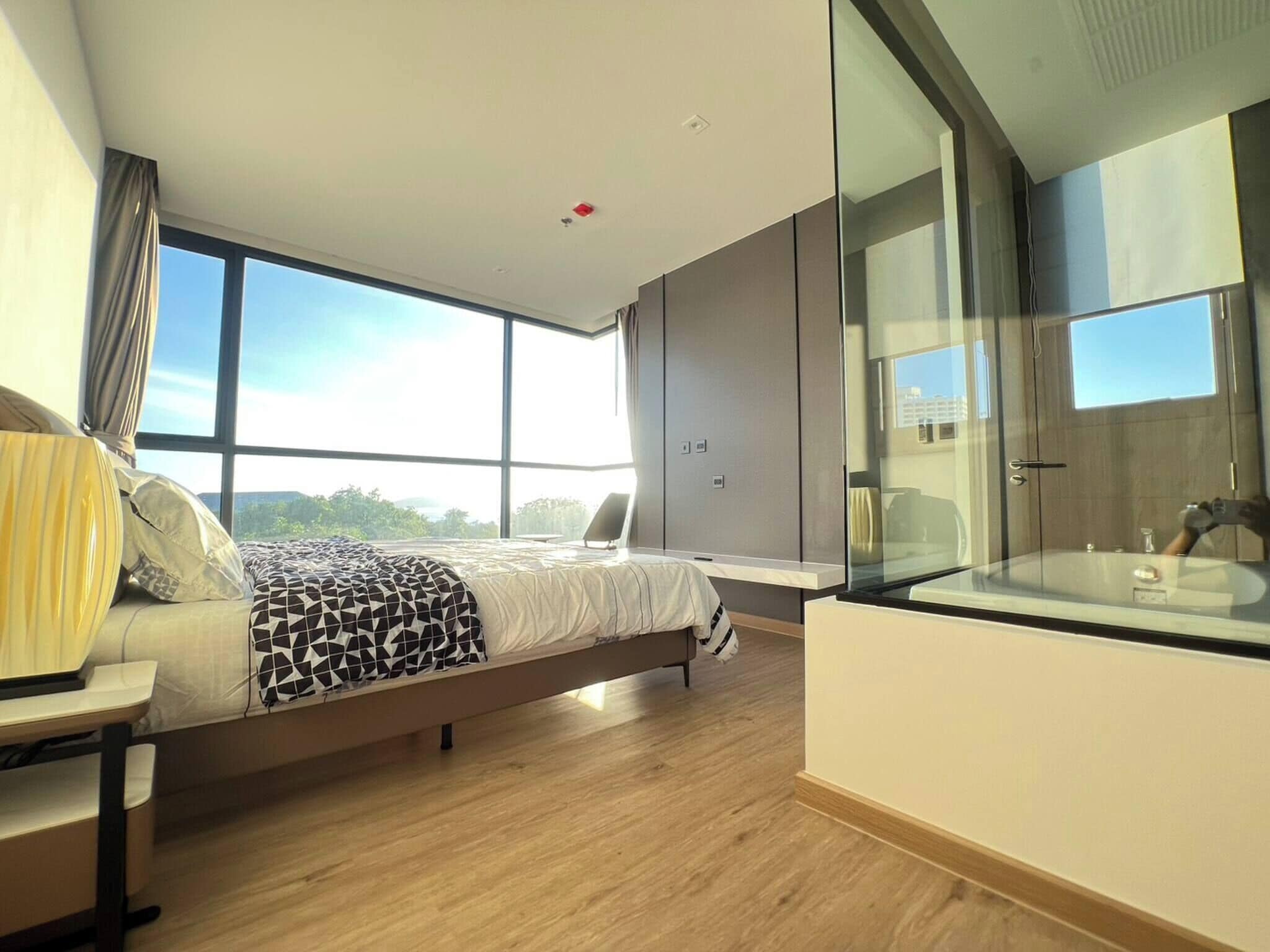Une chambre moderne avec un grand lit, une table de chevet et une salle de bain fermée par des parois de verre, offrant une vue sur l’extérieur.
