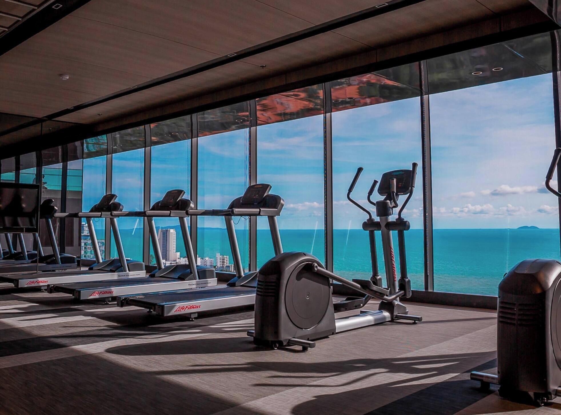 Un gimnasio con cintas de correr, elípticas y una máquina de remo, con vistas al océano y al paisaje urbano a través de amplios ventanales.