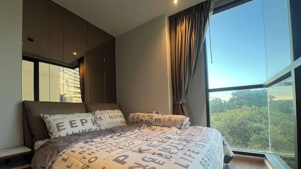 A modern bedroom with a large bed, a view of trees through a window, and a cityscape in the background. - 2 Bed Room Rare Unit With Bath Tub Sea View (Pattaya)