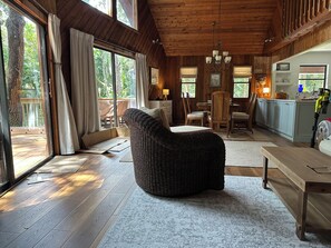 A cozy room with a large window, a wicker chair, a wooden table, and a kitchen area.