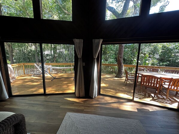 A room with large windows overlooking a wooden deck with a hammock and a table.
