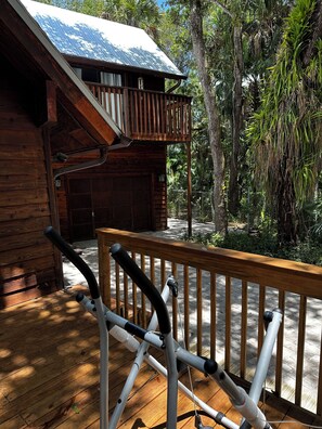 Um deck de madeira com uma bicicleta ergométrica, uma cabana de madeira com telhado metálico e uma floresta verde exuberante ao fundo.