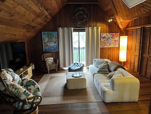 Ein gemütliches Zimmer mit hohen Decken, Holzwänden, einem großen Fenster mit Vorhängen, einer Couch, einem Sessel und Bildern an den Wänden.