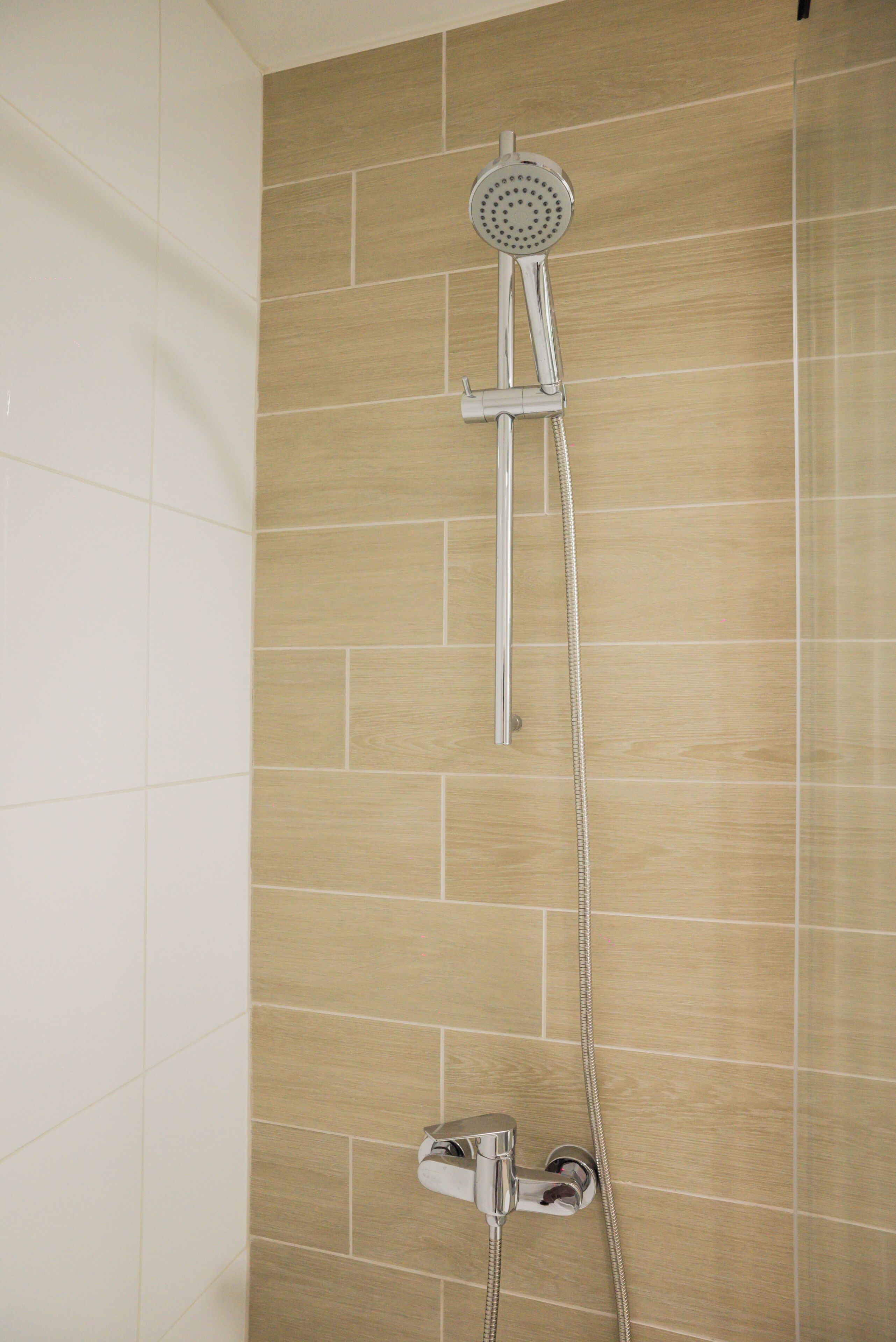 Une douche équipée d’un pommeau de douche à main et d’un pommeau de douche fixe fixé au mur, tous deux dotés de finitions chromées, le tout installé devant un mur recouvert de carreaux de couleur beige.