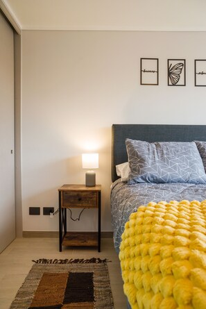 Ein Schlafzimmer mit einem Bett, einem Nachttisch, einer Lampe, Wandkunst und einem gemusterten Teppich.