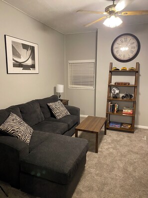 Ein Wohnzimmer mit Ecksofa, Couchtisch, Bücherregal, Wanduhr und Deckenventilator.