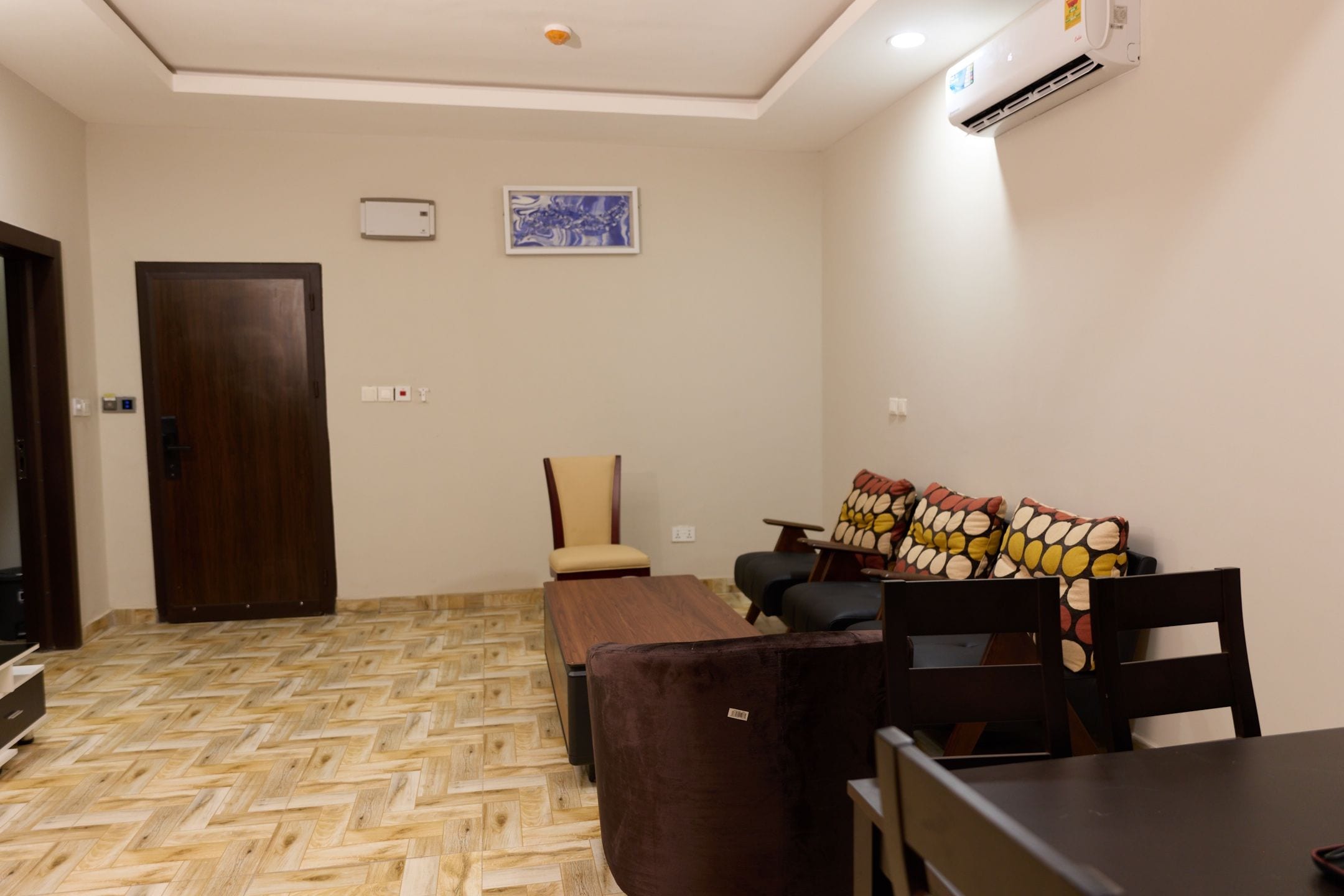 A room with a wooden herringbone floor, a brown sofa with patterned cushions, a wooden chair, a small table, and a door.