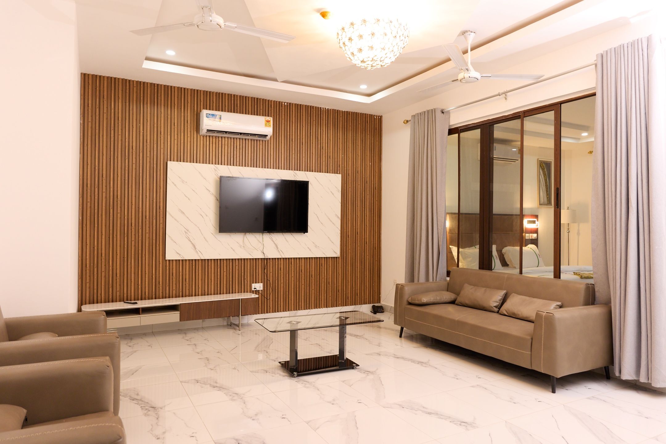 Una sala de estar moderna con un televisor de pantalla plana montado en una pared de paneles de madera, un sofá, una mesa de centro de cristal y un ventanal con cortinas.