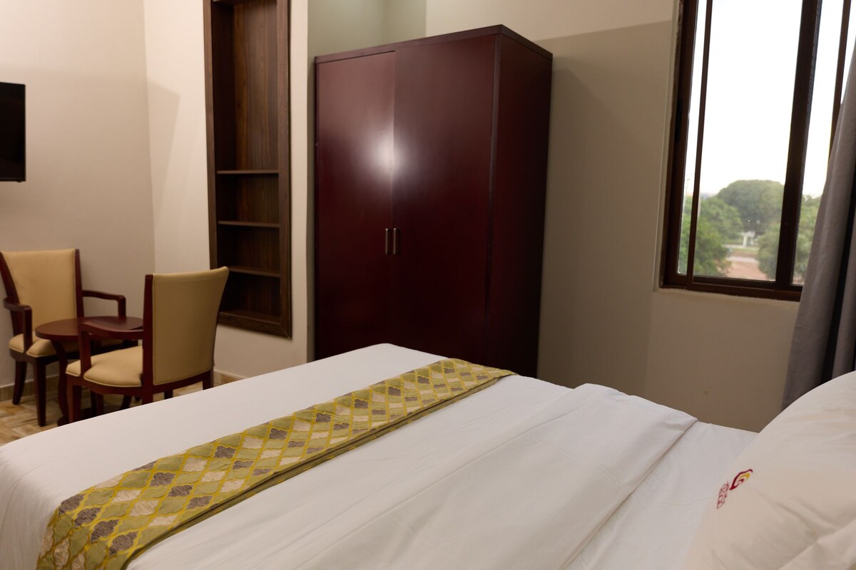 Habitación de hotel con cama, armario, escritorio con sillas y ventana con vistas.