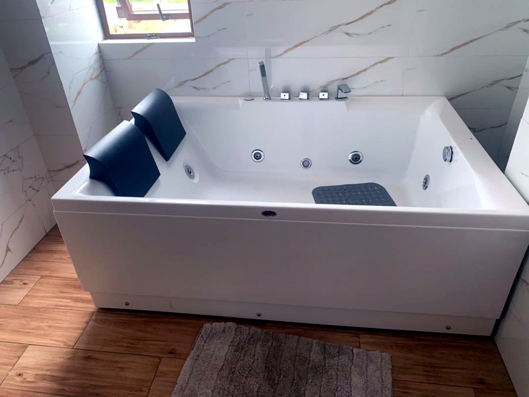 A white freestanding bathtub with a black seat and a small black mat, set against a marble wall with a window above.