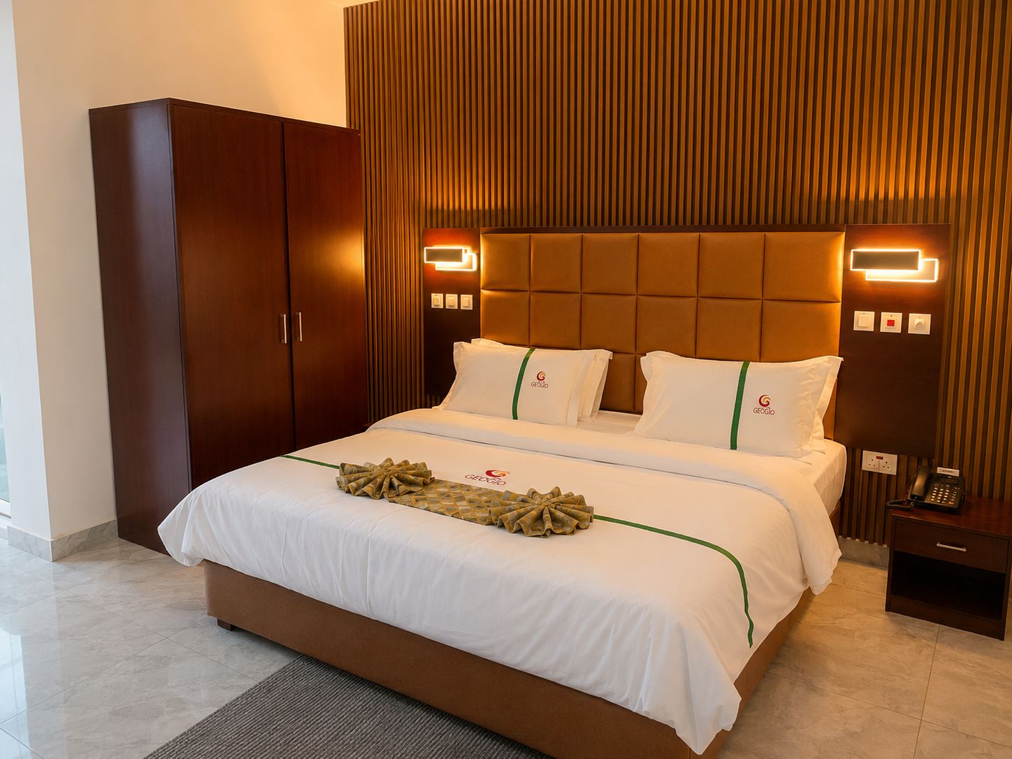 A hotel room with a large bed, a wardrobe, a bedside table with a phone, and a wall-mounted light fixture.