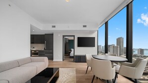 Un salon moderne comprenant un canapé, une table basse et un coin repas avec des chaises, un téléviseur fixé au mur et une vue sur la ville grâce à de grandes fenêtres.