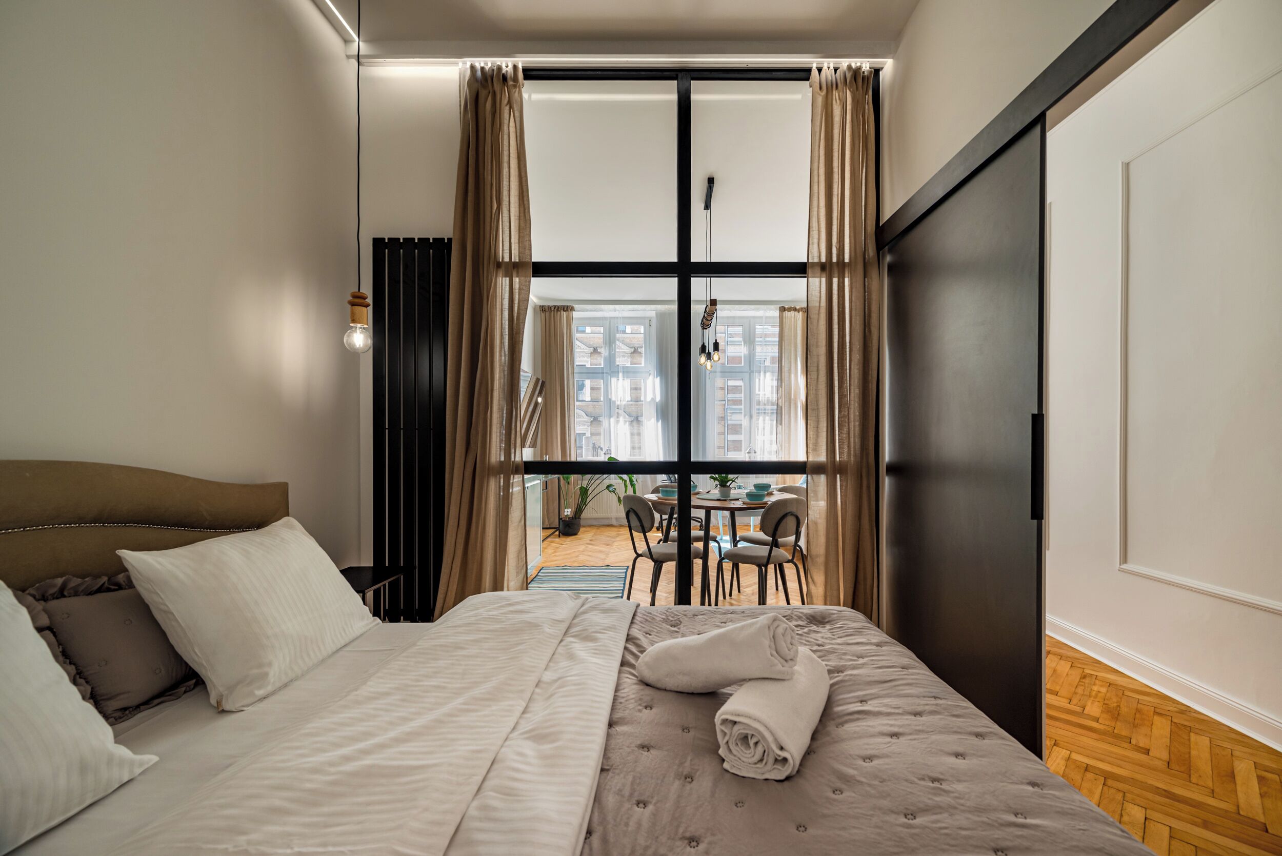 A hotel room with a bed, a dining table with chairs, and a view of the city through large windows.