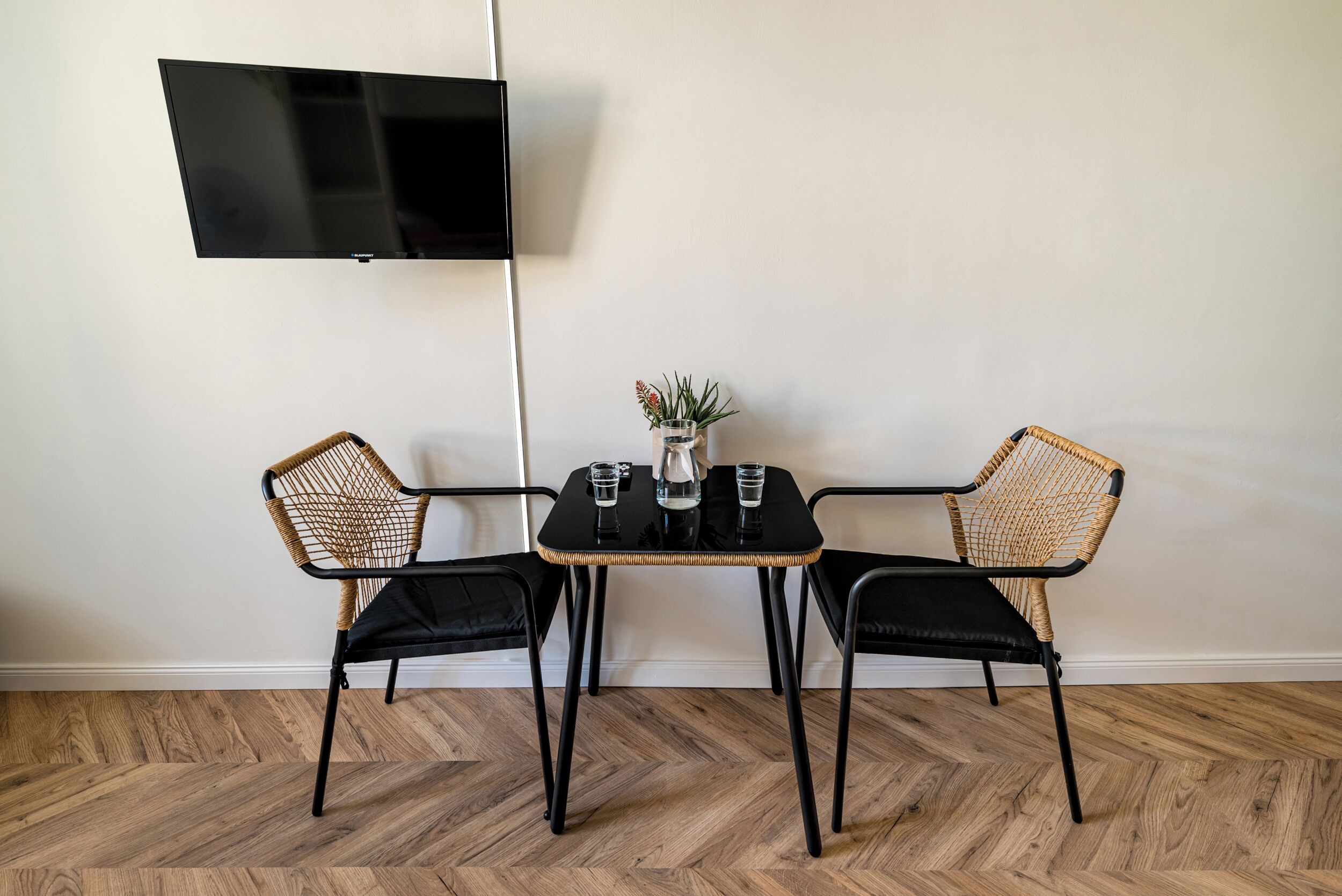Ein kleines Zimmer mit einem schwarzen Tisch und zwei Stühlen mit Geflochtensitzen, einem an der Wand montierten Flachbildfernseher und einem Blumentopf auf dem Tisch.